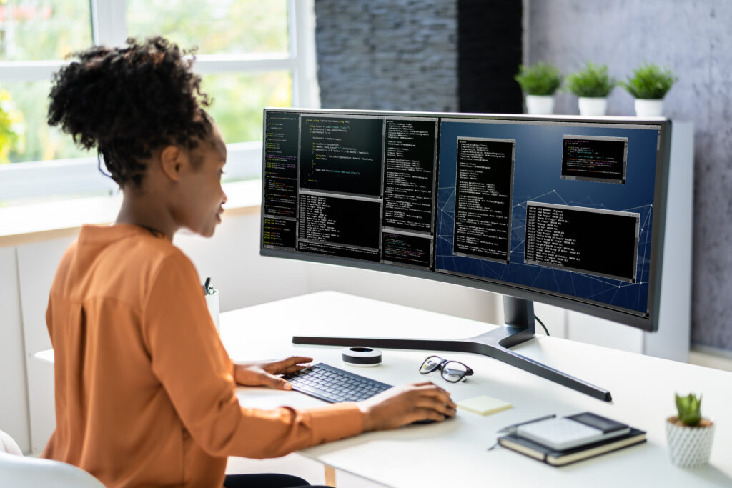 Black female software developer seated at a desk looking at a dual monitor.