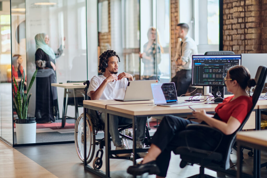A young business group, including an African American businessman in a wheelchair, collaborates within a modern glass office, actively engaged around a computer and laptop, collectively solving diverse business challenges with determination.